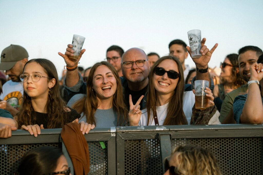 Gruppe fröhlicher Konzertbesucher an der Absperrung, einige halten Plastikbecher mit Bier; eine Frau macht ein Peace-Zeichen.