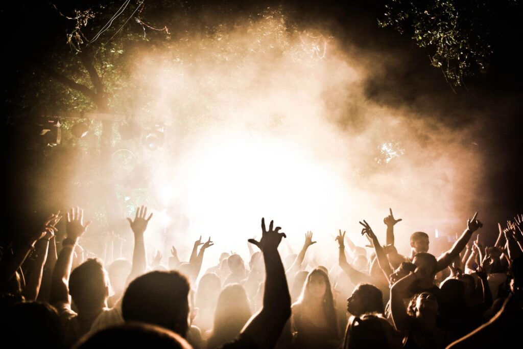 Dunkle Menschenmenge bei einem Konzert, viele Personen heben die Hände vor starker Bühnenbeleuchtung und Nebel.