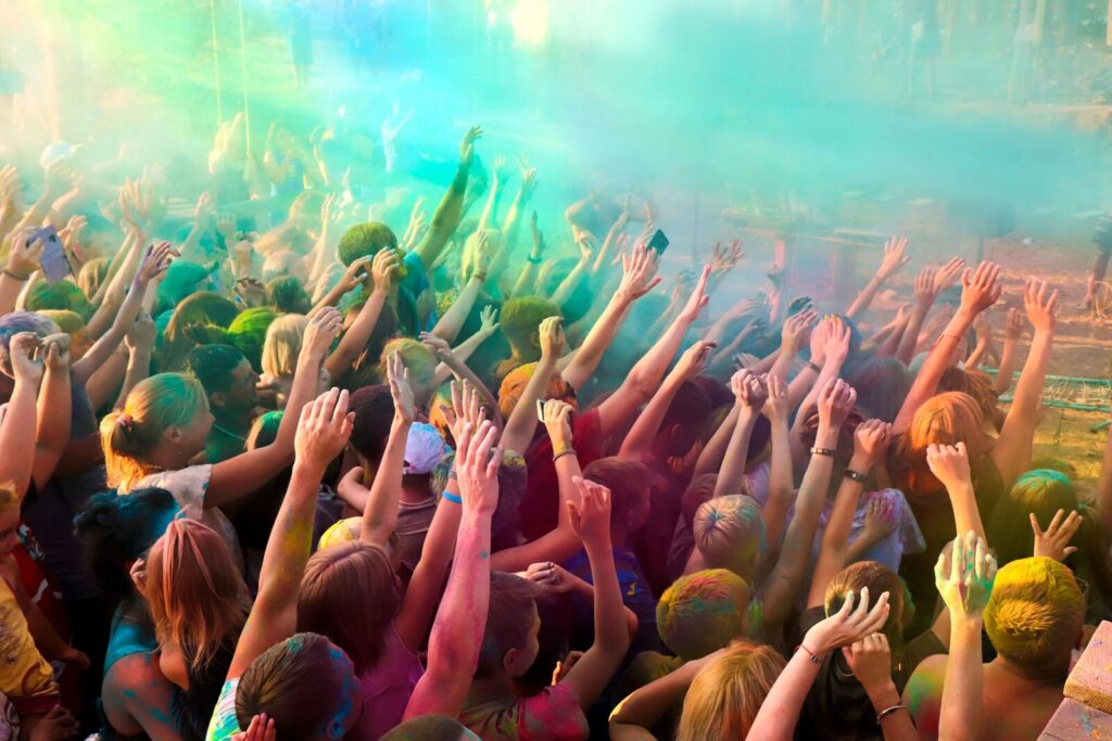 Menschenmenge bei einem Farbfestival, viele Teilnehmende mit erhobenen Händen und bunter Farbe auf Haut und Kleidung.