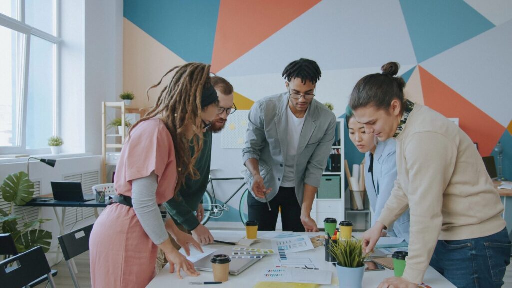 Team von fünf Personen berät sich an einem Tisch mit Dokumenten, Farbkarten und Kaffeebechern in einem modernen Büro.